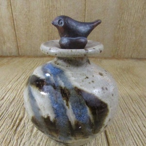 Puede incluir: Un tarro de cerámica con una tapa en forma de pájaro. El tarro está decorado con esmalte marrón, azul y blanco. El pájaro es marrón y tiene un ojo negro.