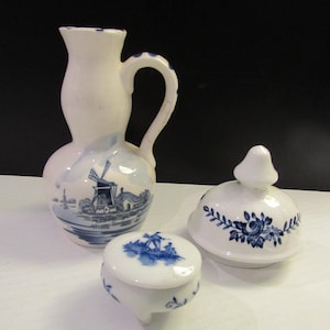 May include: A set of three blue and white ceramic pieces with a Dutch windmill design. The set includes a pitcher, a lidded jar, and a small round dish with three legs.