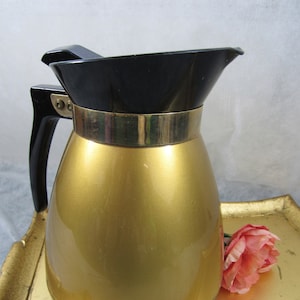 May include: A gold-coloured thermal carafe with a black lid and a gold band around the top. The carafe is sitting on a gold-coloured tray.