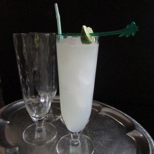 May include: A tall glass filled with a white, cloudy drink with ice. A lime wedge and a green plastic palm tree straw are in the glass. The glass is on a silver tray with another empty glass.