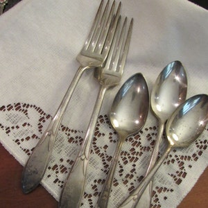 May include: Four silver forks and three silver spoons on a white lace tablecloth. The forks are stacked on top of each other, and the spoons are arranged in a fan shape.