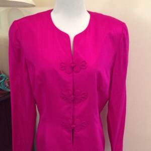 Op de afbeelding: Een levendig fuchsia tweedelig pak op een mannequin. De jas heeft een mandarijnkraag en decoratieve bloemvormige knopen. De bijpassende rok is een rechte, knielange stijl, wat zorgt voor een gepolijste en elegante ensemble.