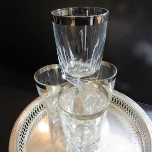 May include: Four clear glass tumblers with silver rims sit on a silver tray. The tumblers are stacked on top of each other.