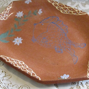 May include: A terracotta-colored ceramic serving dish with a blue fish design and white floral accents. The dish has a scalloped edge with a white and yellow design.