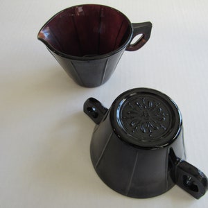 May include: A set of two vintage glass creamer and sugar bowls. The creamer is a deep purple color and the sugar bowl is black. Both have a fluted design and a decorative floral pattern on the bottom.