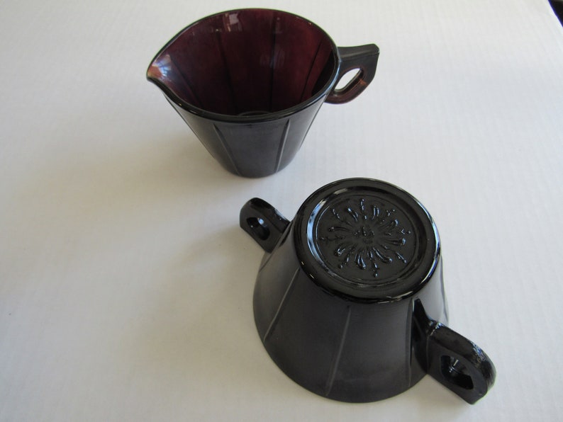 May include: A set of two vintage glass creamer and sugar bowls. The creamer is a deep purple color and the sugar bowl is black. Both have a fluted design and a decorative floral pattern on the bottom.