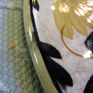 May include: A section of a decorative plate with a floral design. The plate has a white background with a cracked glaze effect. Black leaf shapes and yellow flower petals are visible, along with a thin brown stem.