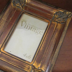 K&ouml;nnte beinhalten: Ein brauner und goldener Bilderrahmen mit verzierten Blattdetails und einer goldenen Inschrift mit dem Text "Philip Whitney Ltd. Vintage 4x6".