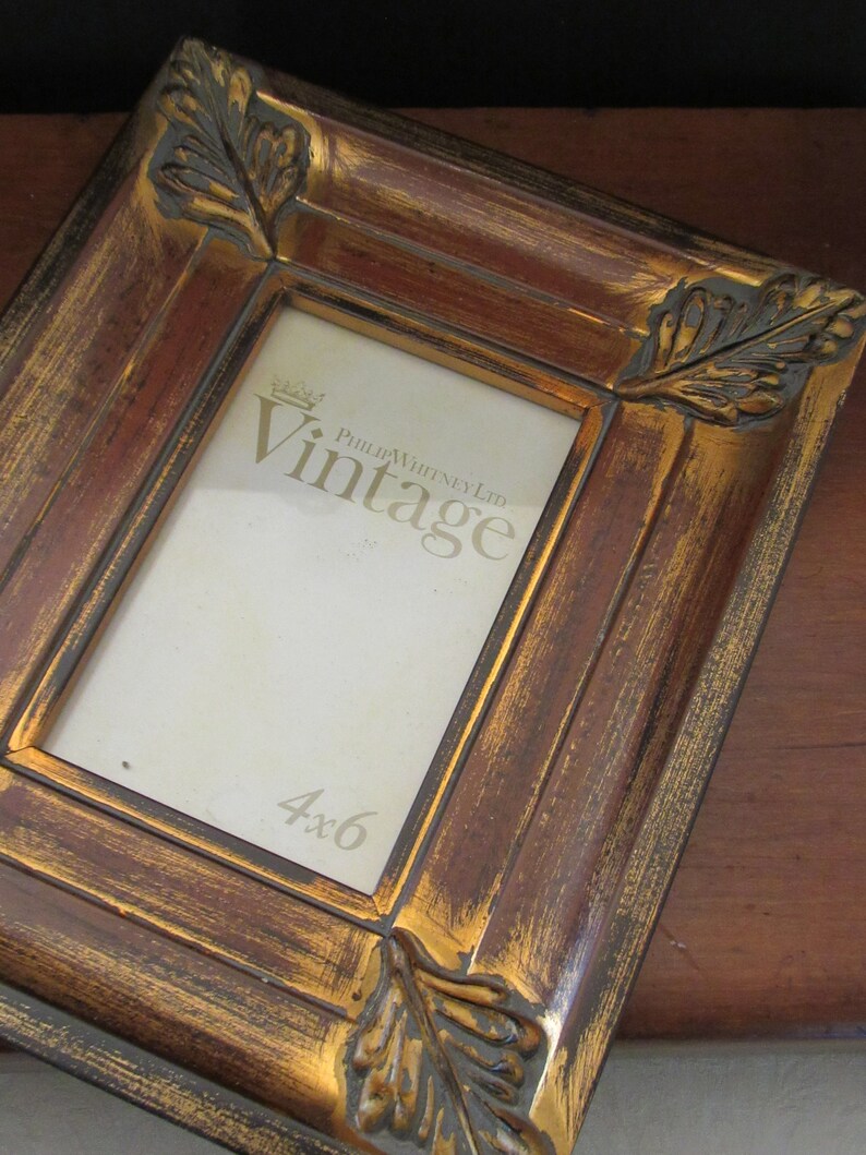 K&ouml;nnte beinhalten: Ein brauner und goldener Bilderrahmen mit verzierten Blattdetails und einer goldenen Inschrift mit dem Text "Philip Whitney Ltd. Vintage 4x6".