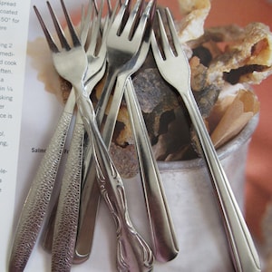 May include: A collection of five silver-toned metal forks with varying handle designs, including textured, patterned, and smooth finishes, arranged in a cluster. The forks feature three tines each and are made of polished stainless steel material.