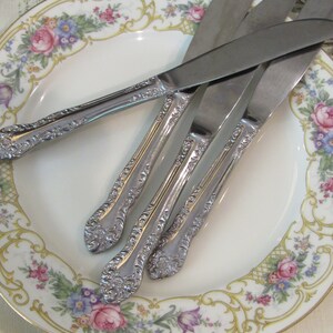 May include: Four silver dinner knives with ornate handles are arranged on a white plate with a floral pattern. The plate has a gold rim and a floral design with pink, yellow, and green flowers.