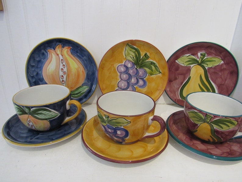 May include: Set of six ceramic teacups and saucers with fruit designs. The cups and saucers feature a variety of fruits, including pears, grapes, and pomegranates. The designs are painted in bright colors on a white background.