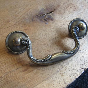May include: Antique brass drawer pull with a decorative rope-like border around the circular backplates. The curved handle features a beaded design. The hardware is set against a light-colored wooden surface.