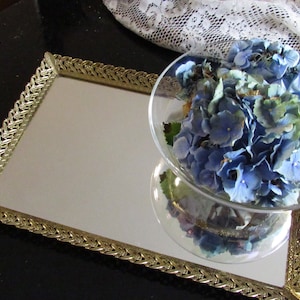 May include: A gold-coloured, ornate, rectangular tray with a mirrored surface. A glass bowl filled with blue hydrangeas sits on the tray.