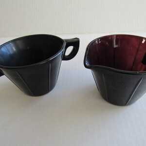 May include: A set of two black glass creamer and sugar bowls with fluted sides and two handles. The creamer is a darker shade of purple.
