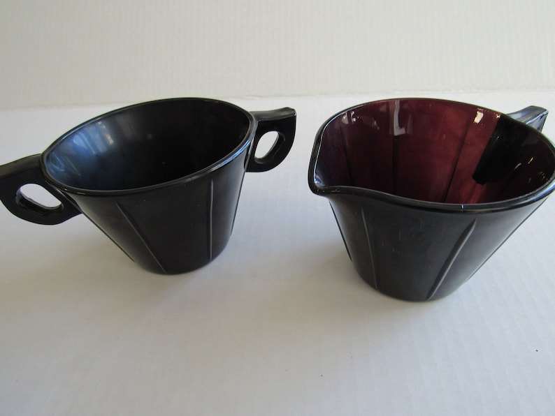 May include: A set of two black glass creamer and sugar bowls with fluted sides and two handles. The creamer is a darker shade of purple.