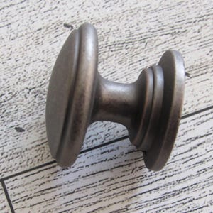 May include: A bronze-colored cabinet knob with an oval face and a cylindrical base. The knob has a slightly aged appearance, suggesting a vintage or antique style. The hardware is designed for use on drawers or cabinet doors.