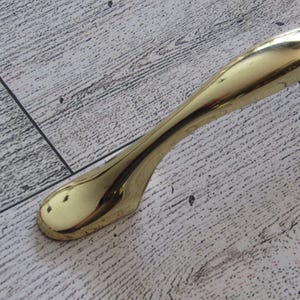 May include: A curved, polished brass door handle. The handle has a smooth, ergonomic design and a bright, reflective surface. The handle is likely for a cabinet or drawer, and is shown against a light wood grain background.