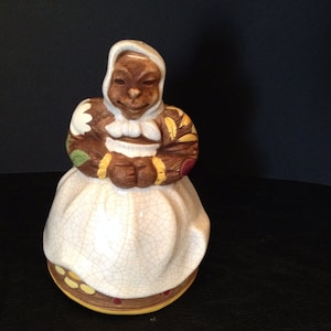 Puede incluir: Figura de cerámica de una mujer sentada con un gorro y un vestido blancos. La figura tiene piel morena, una expresión sonriente y detalles marrones, amarillos y verdes. La base está decorada con círculos amarillos y rojos.