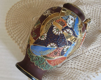 May include: A brown ceramic vase with a gold, blue, red and white painted design of a person in traditional Japanese clothing. The vase has a round body and a narrow neck.