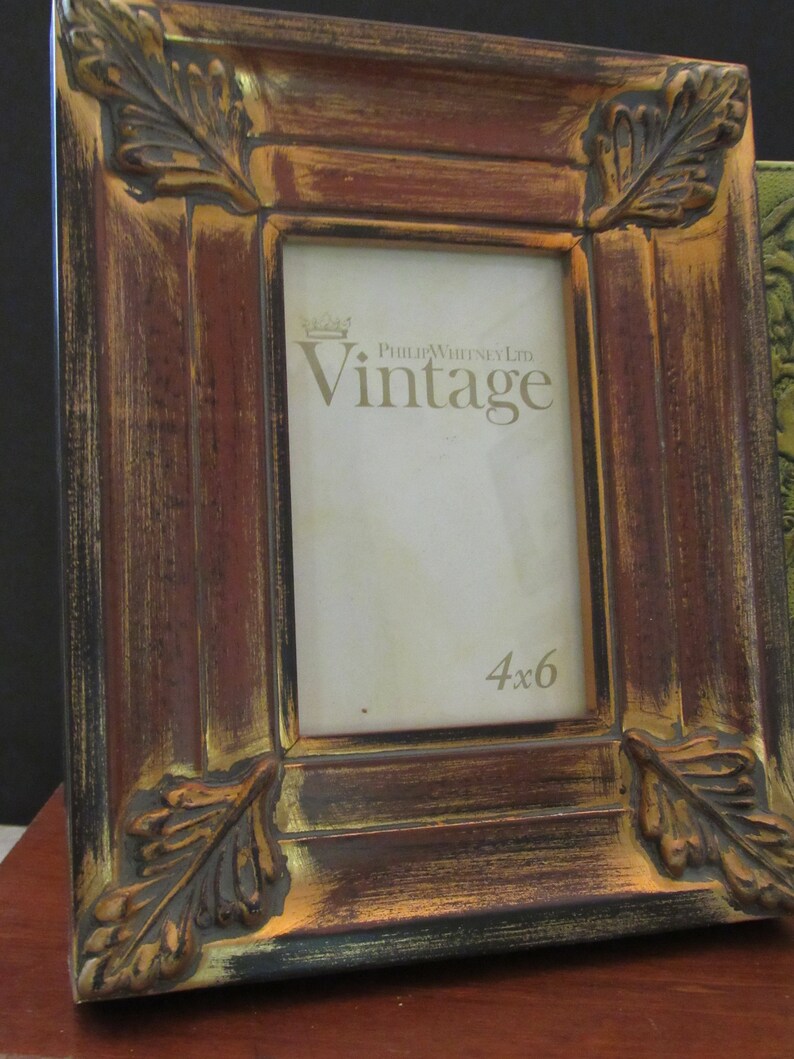 K&ouml;nnte beinhalten: Ein verwitterter Holzbilderrahmen mit einer Blattgold-Oberfl&auml;che. Der Rahmen hat an jeder Ecke ein erhabenes Blattdesign und tr&auml;gt die Aufschrift "Philip Whitney Ltd. Vintage" und "4x6" auf der Vorderseite.