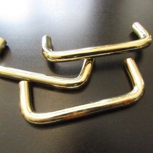 May include: Three gold-toned metal cabinet door pulls. Each pull is shaped like a rounded rectangle with a slight curve at the top.