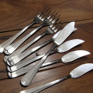 Puede incluir: Un conjunto de cubiertos antiguos, que incluye tenedores y cuchillos de mantequilla, dispuestos sobre una superficie de madera. Los tenedores tienen cuatro púas y los cuchillos de mantequilla tienen una hoja curva. Los cubiertos son de metal brillante color plata.