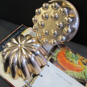 May include: Two vintage copper cake molds. One mold is round with a raised design of 16 circles. The other mold is a flower shape with 10 petals. The molds are sitting on a cookbook open to a page titled "Fruit Salads".
