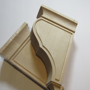 May include: Two unfinished wooden corbels, ready for staining or painting. The corbels have a decorative, curved design with a rectangular panel. They are wrapped in clear plastic, suggesting they are new and ready for use in home decor projects.