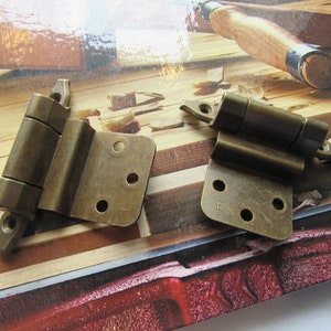 May include: Two brass hinges with a satin finish. The hinges are on a wooden surface.