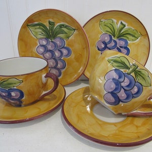 May include: A set of four yellow ceramic teacups and saucers with a grape design. The cups and saucers have a brown rim and are decorated with purple grapes, green leaves, and a yellow background.