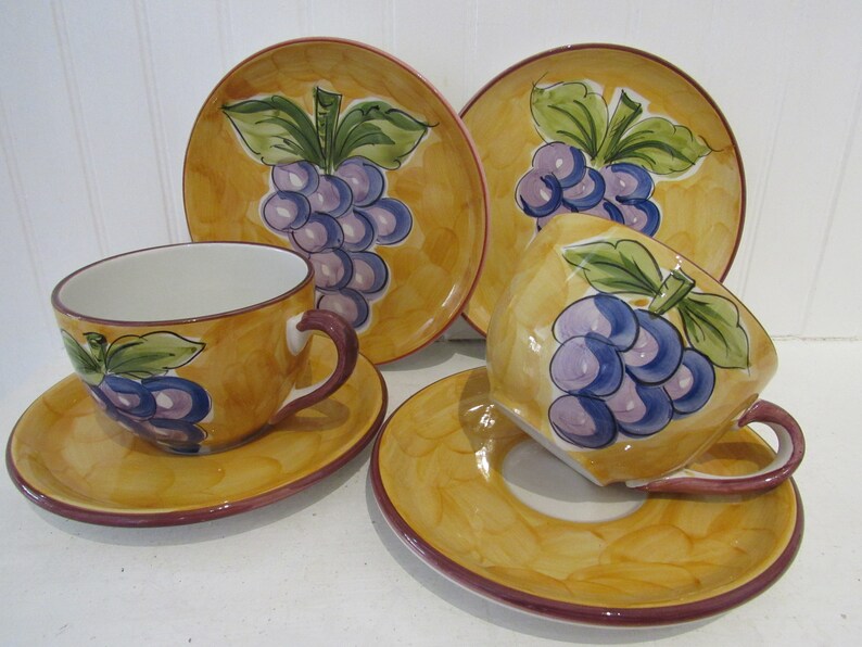 May include: A set of four yellow ceramic teacups and saucers with a grape design. The cups and saucers have a brown rim and are decorated with purple grapes, green leaves, and a yellow background.
