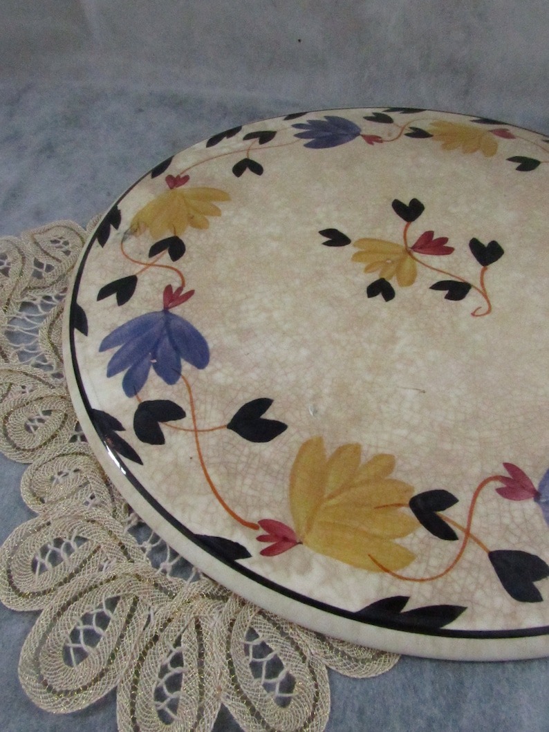 May include: Oval ceramic serving platter with a cream-colored background and a floral design. The flowers are yellow, blue, and red, with black stems and leaves. The edge of the platter is black.