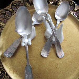 Puede incluir: Una colección de cucharas de plata antiguas exhibidas en un plato decorativo dorado. Las cucharas están sostenidas por soportes de cerámica blanca con forma de mano. Las cucharas tienen un aspecto vintage y deslustrado.