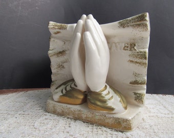May include: White ceramic bookends with gold accents depicting two hands clasped in prayer. The bookends are on a white lace surface. The text "PRAYER" is visible on the bookends.