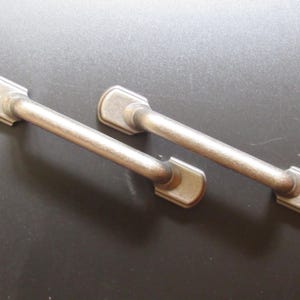 May include: Two antique-style cabinet pulls with a brushed metal finish. Each pull features a rectangular base with rounded edges and a straight, cylindrical handle. The hardware is designed for furniture and home decor projects.