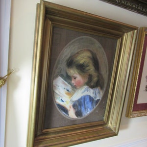 May include: A framed painting of a young girl in a blue dress reading a book. The painting is set against a brown background and is framed in a gold frame.
