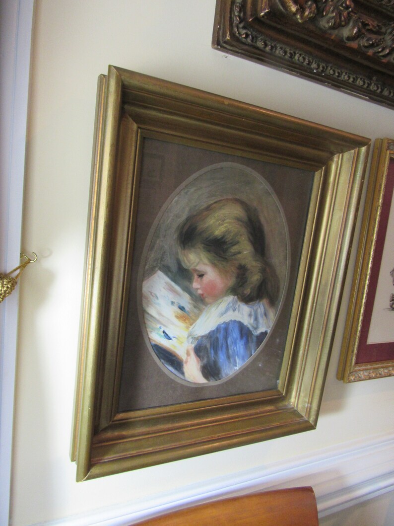 May include: A framed painting of a young girl in a blue dress reading a book. The painting is set against a brown background and is framed in a gold frame.