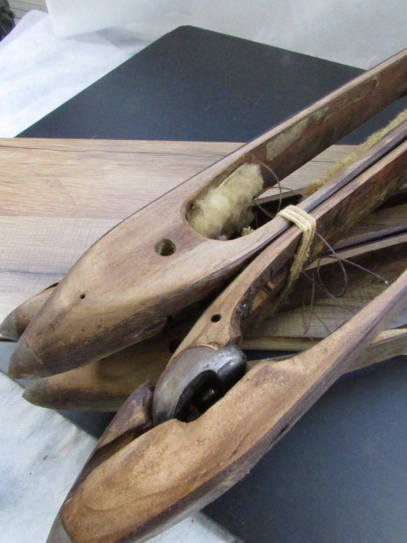 May include: Three wooden weaving shuttles with a string tied around them. The shuttles are made of light brown wood and have a hole in the centre. One shuttle has a white cotton filling inside.