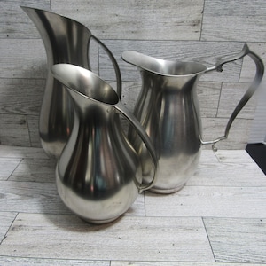 May include: Three silver pitchers of varying sizes and shapes on a light wood background.