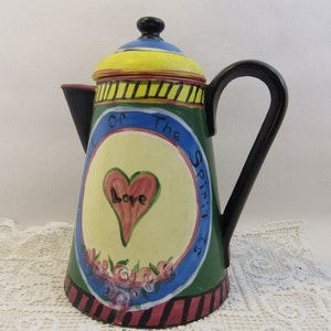 Puede incluir: Una cafetera de cerámica verde, amarilla, azul y roja con asa negra. La cafetera tiene un corazón con la palabra "Love" en su interior y el texto "Fruit of the Spirit is" alrededor.