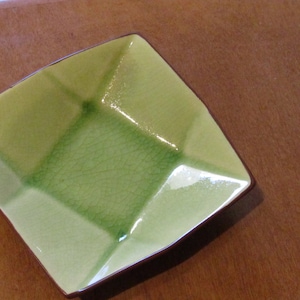 Puede incluir: Un plato cuadrado de cerámica verde con un borde marrón. El plato tiene un esmalte craquelado y un centro ligeramente elevado.