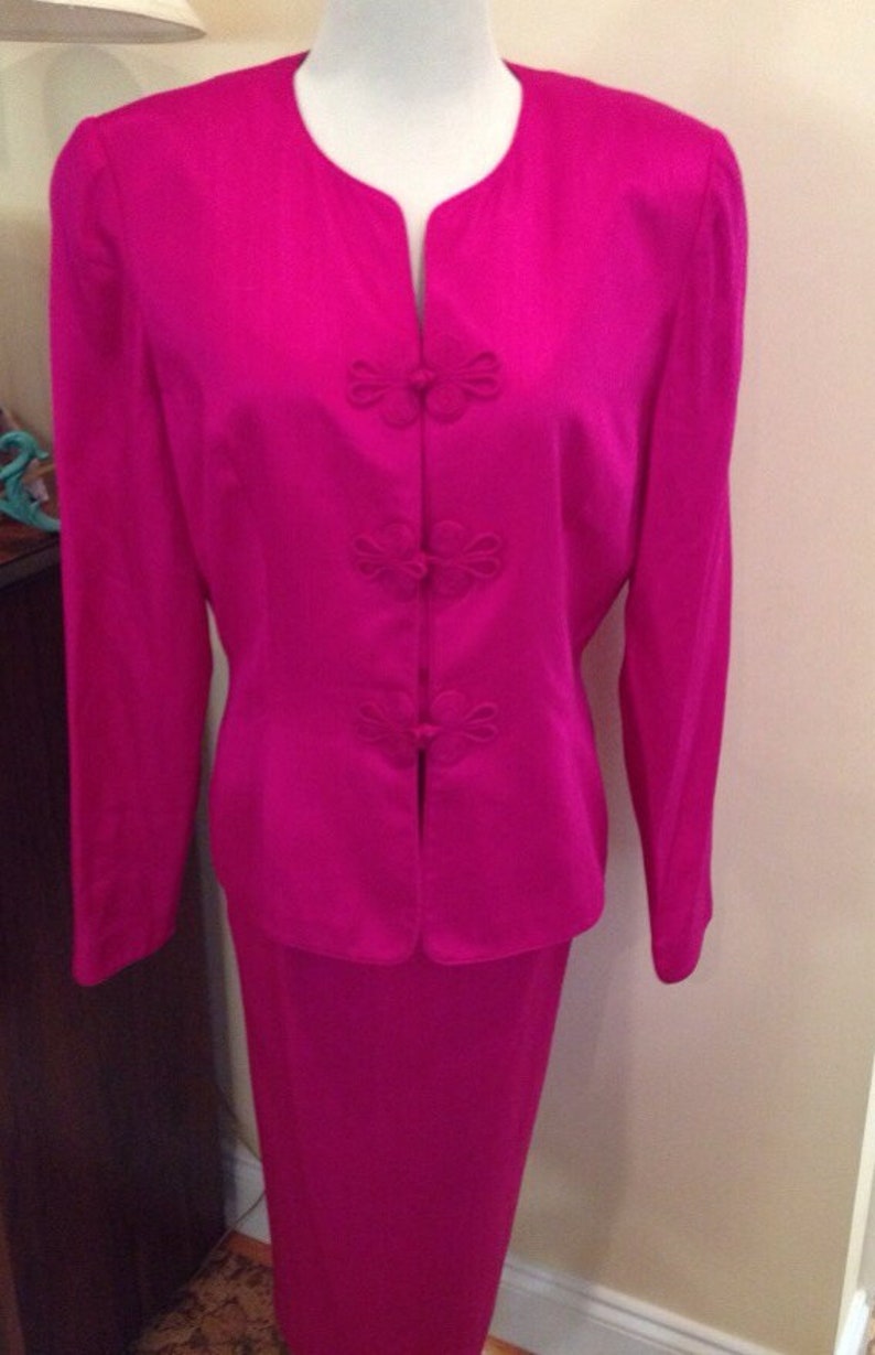 Op de afbeelding: Een levendig fuchsia tweedelig pak op een mannequin. De jas heeft een mandarijnkraag en decoratieve bloemvormige knopen. De bijpassende rok is een rechte, knielange stijl, wat zorgt voor een gepolijste en elegante ensemble.