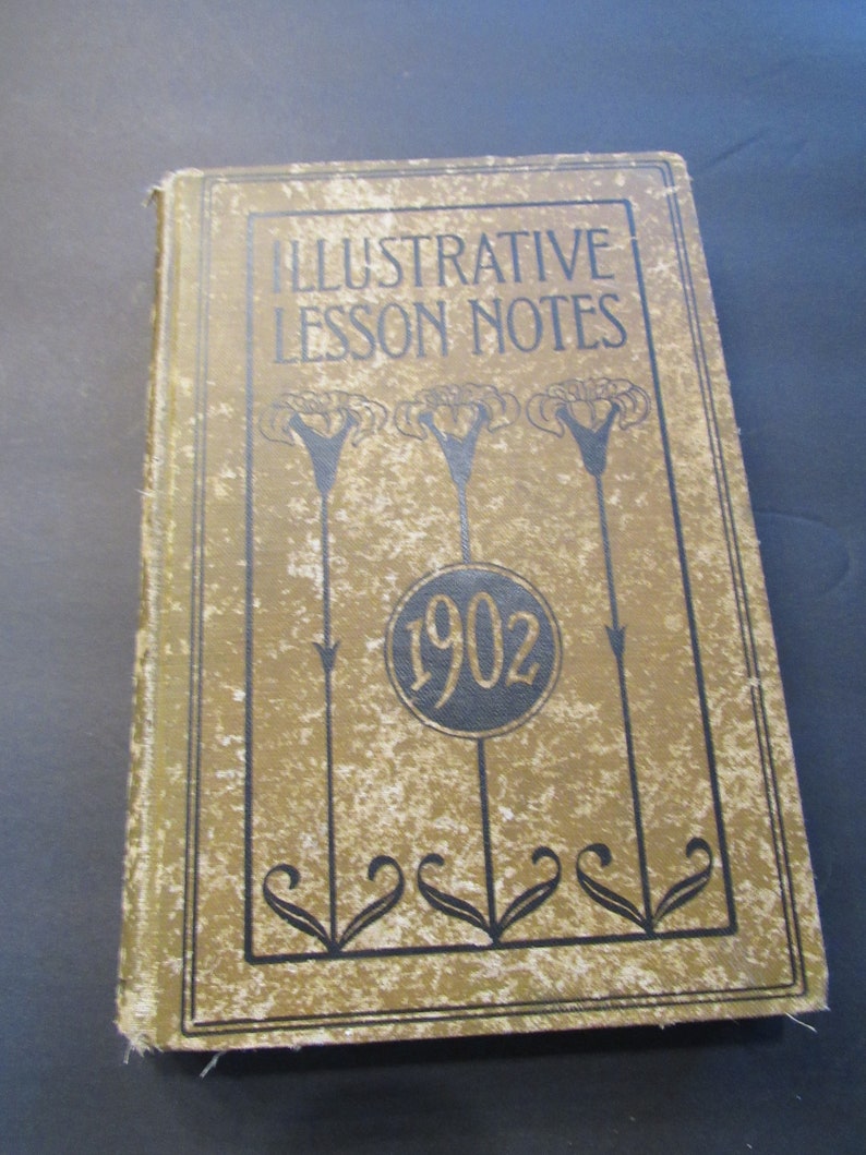 Pu&ograve; includere: Un libro vintage intitolato "Illustrative Lesson Notes" con una copertina marrone e testo nero. Il libro presenta un design decorativo con fiori stilizzati e un cerchio con l'anno "1902" stampato su di esso.