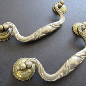 May include: Two ornate brass cabinet door pulls with a decorative, swirling design. The pulls have a round base and a curved handle.