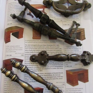 May include: A collection of antique brass door and drawer pulls. The pulls are in various styles, including a ring pull, a bail pull, and a knob pull. The pulls are all in a worn brass finish.