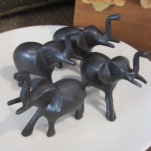 May include: A set of four dark grey elephant figurines, each depicted in a standing position with trunks raised upwards. The smooth, metallic-looking statues are arranged in a cluster, showcasing detailed ears, tusks, and legs.