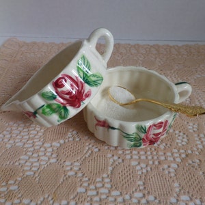 May include: Two white ceramic creamer or gravy boats with a floral design featuring pink roses and green leaves. The boats are on a white crocheted doily.
