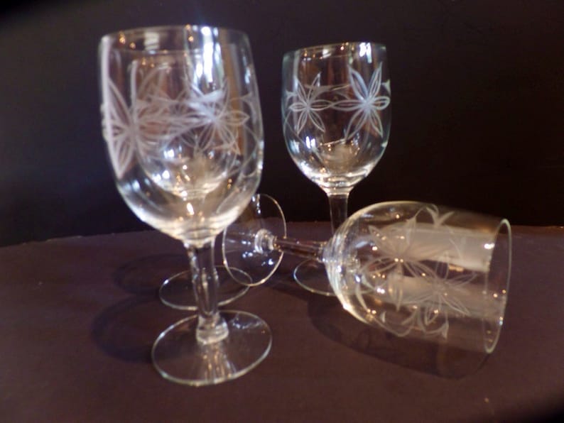 May include: Four clear glass wine glasses with etched floral designs. The glasses are arranged in a group on a dark surface.