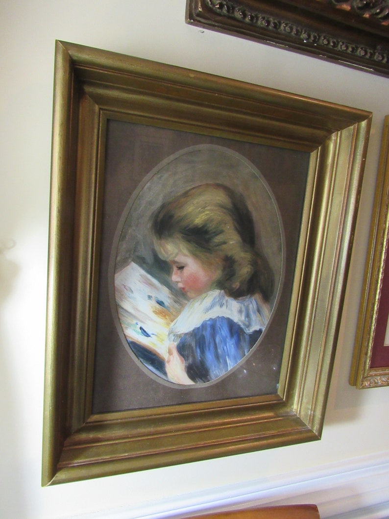 May include: A framed painting of a young girl in a blue dress reading a book. The painting is set against a brown background and is framed in a gold frame.
